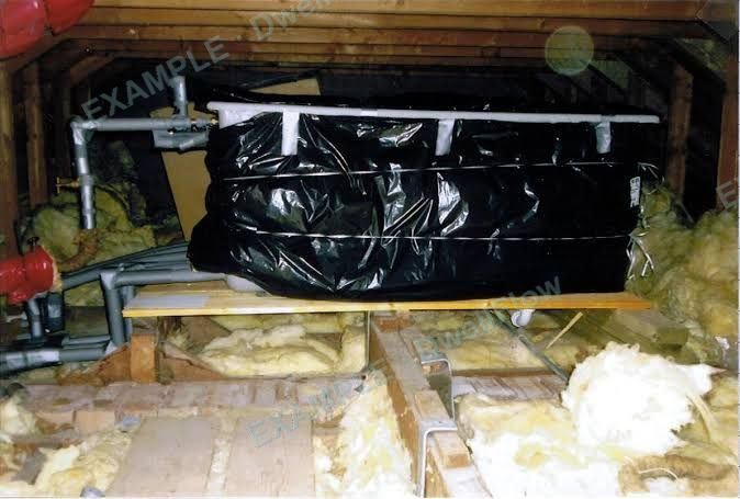 A cold water storage tank wrapped in a black insulating jacket, standing on boards in a loft